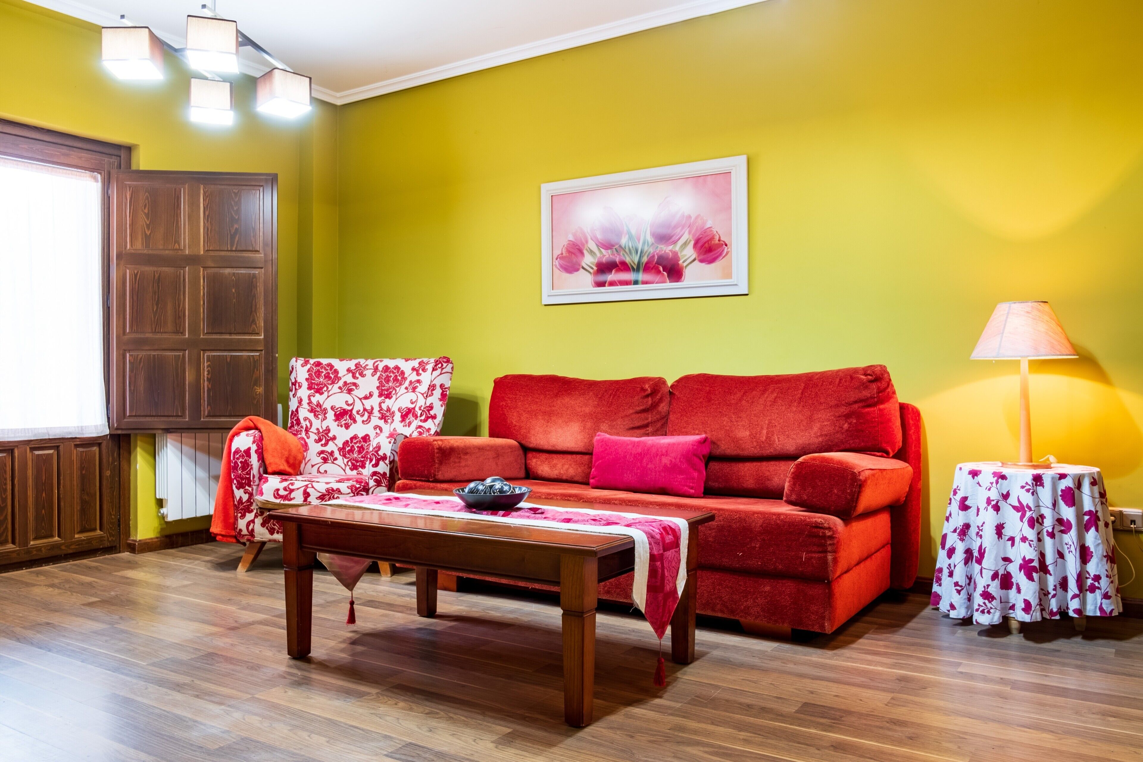 Una sala de estar con un sofá rojo, una mesa de centro de madera, un sillón estampado, una mesita redonda con una lámpara y un cuadro en la pared.