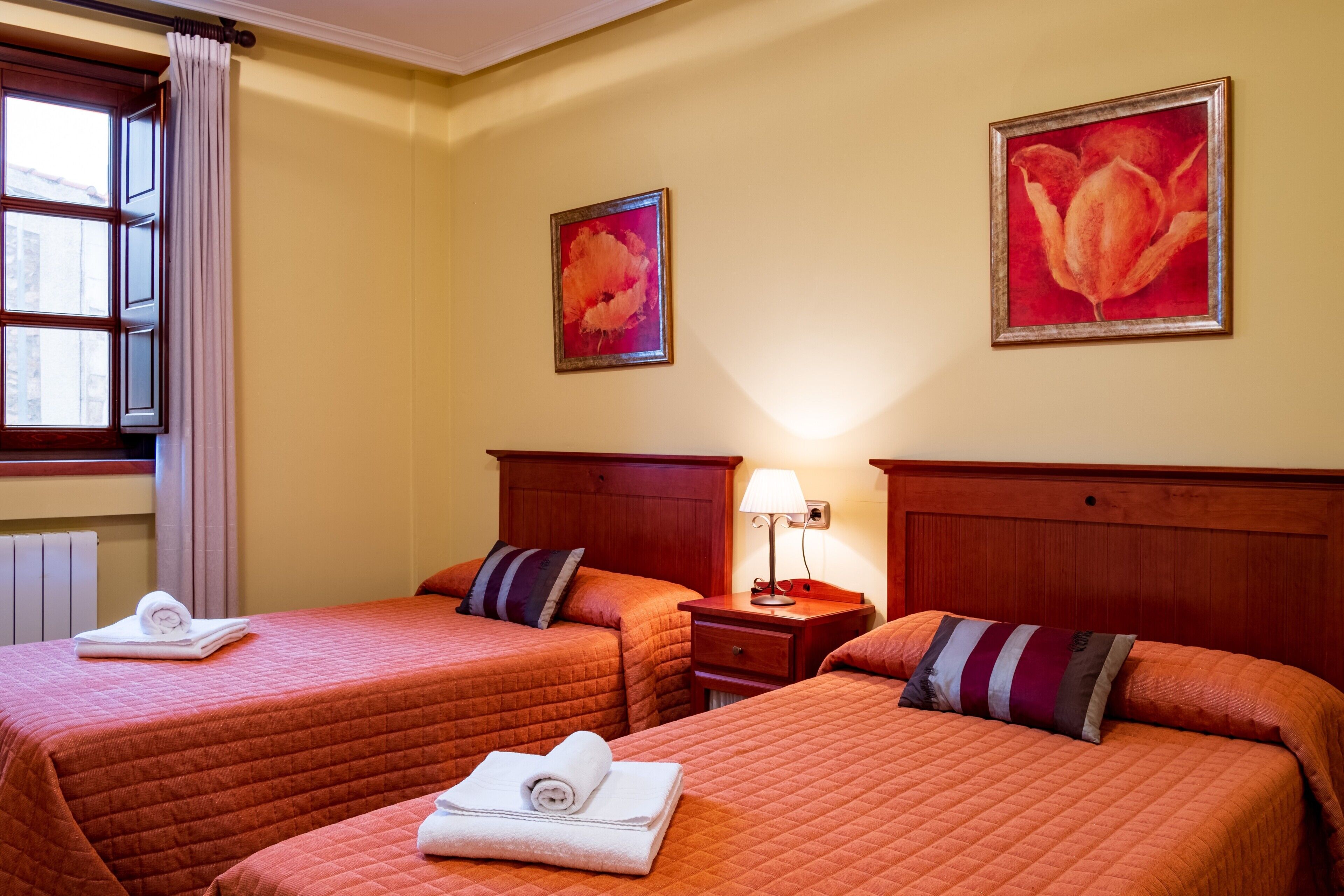 Ein Hotelzimmer mit zwei Betten, einem Fenster mit Vorhängen, einem Nachttisch mit Lampe und zwei Bildern an der Wand.