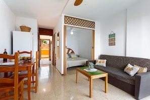 A modern living room with a sofa, a dining table with chairs, a kitchen area with a refrigerator, and a bedroom with a bed and a window.