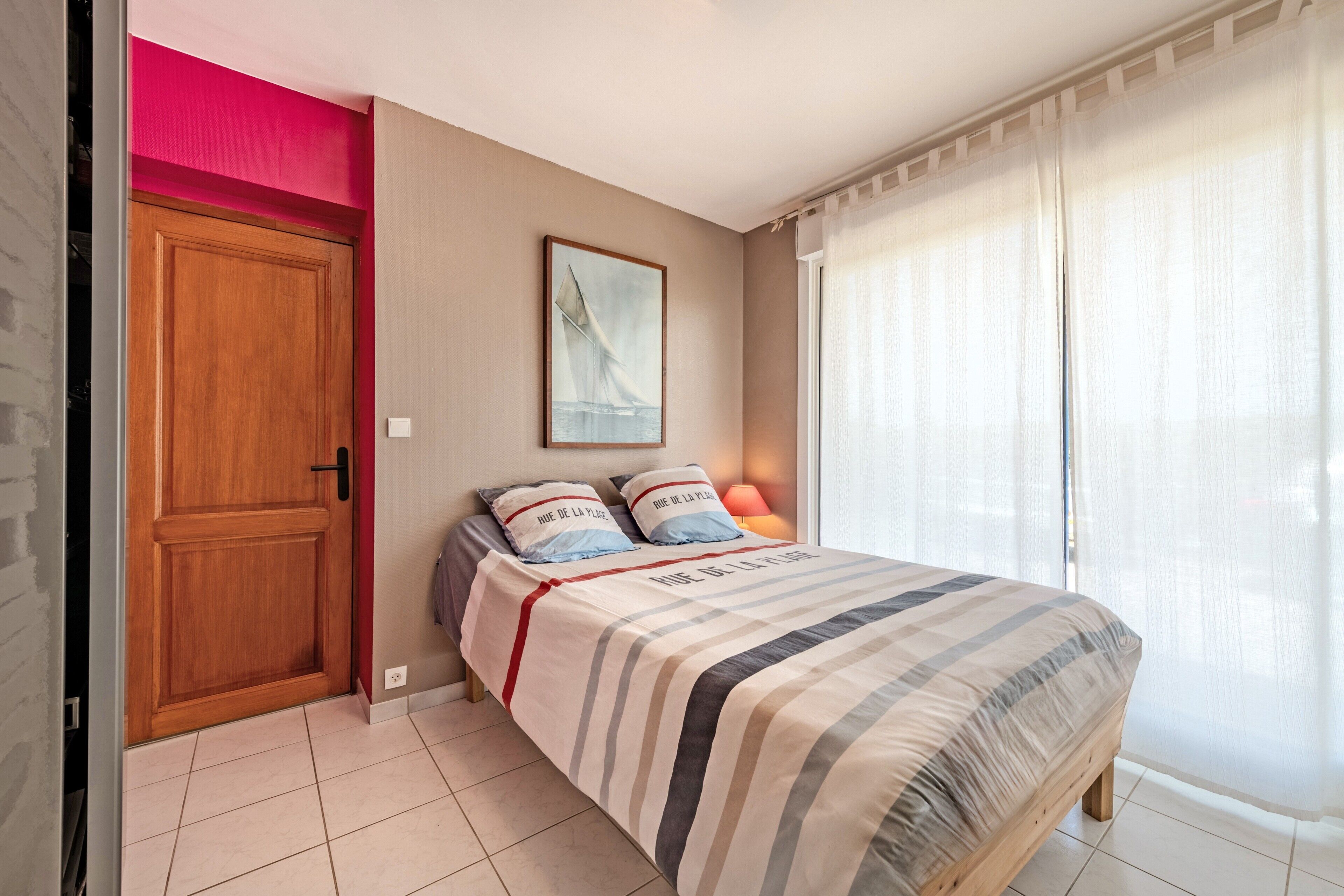 Une chambre à coucher avec un lit, une porte, une fenêtre avec des rideaux, un tableau au mur et une lampe.