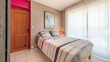 Une chambre à coucher avec un lit, une porte, une fenêtre avec des rideaux, un tableau au mur et une lampe.