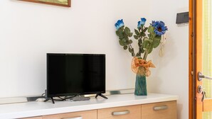 Un téléviseur à écran plat posé sur un comptoir blanc, un meuble en bois avec des tiroirs et un vase avec des fleurs accroché au mur.