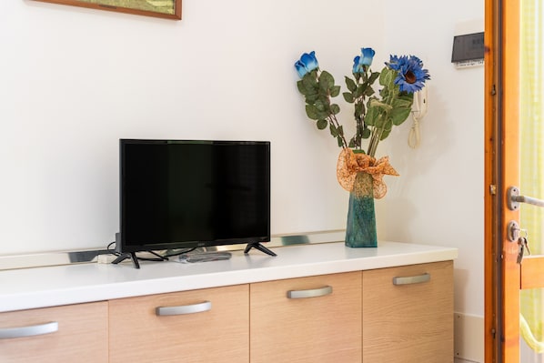 Et fladskærms-tv på en hvid bordplade, et træskab med skuffer, og en vase med blomster på væggen.
