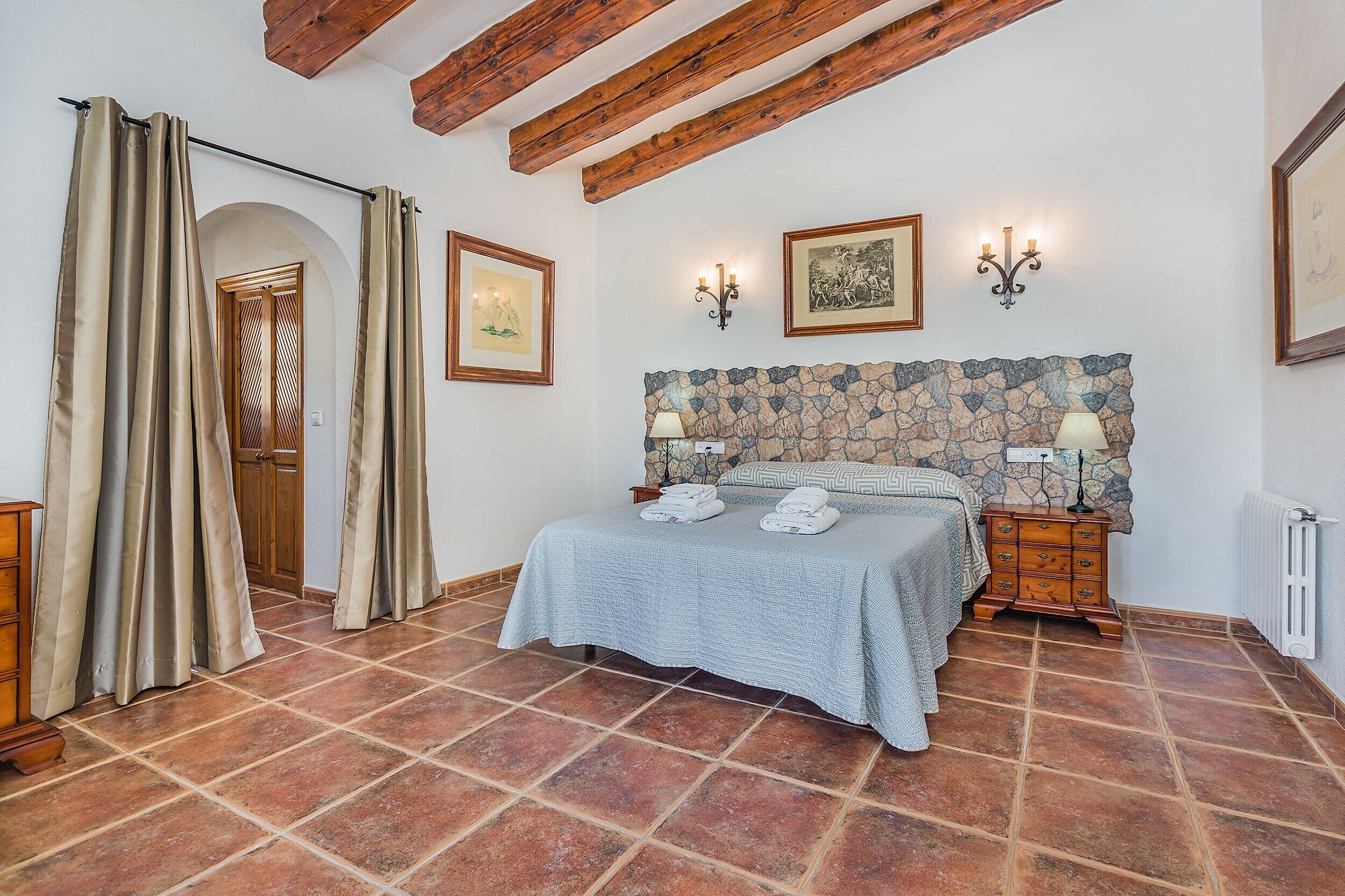 A bedroom with a stone headboard, a bed with towels, a wooden nightstand, and a large window with curtains.