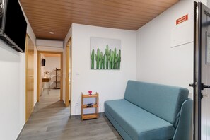 Huoneessa on turkoosinsininen sohva, puinen hylly, jossa on maljakko, kaktusaiheinen taulu ja ovi, jossa on kyltti.