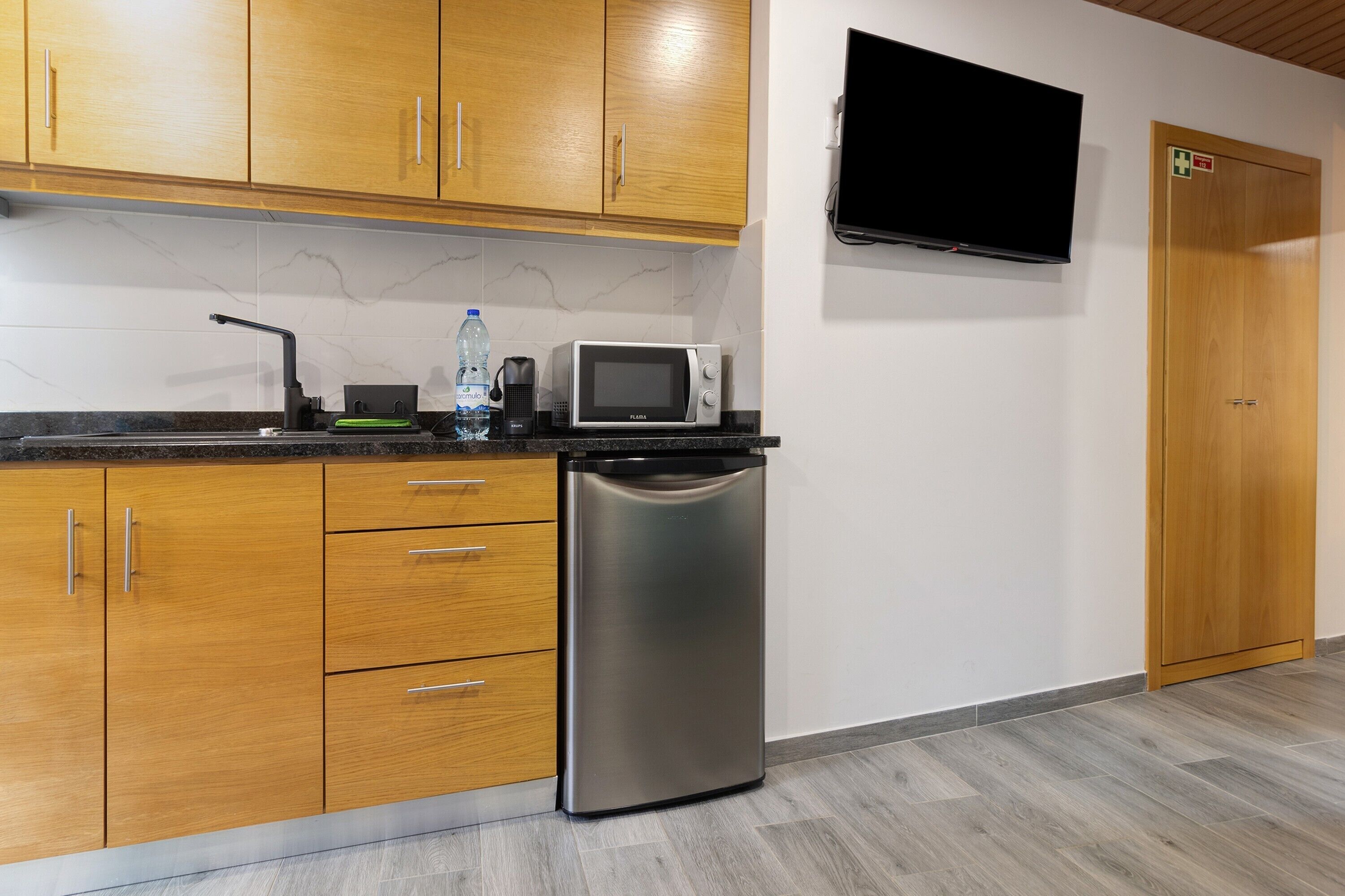 A kitchen with wooden cabinets, a stainless steel refrigerator, a microwave, a water dispenser, and a flat-screen TV mounted on the wall.