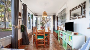 A dining area with a wooden table and chairs, a hanging light fixture, a television on a stand, and a window with curtains. - Villa 'El Limonar' with Shared Pool and Wi-Fi (Chiclana de la Frontera)