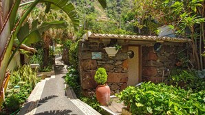 Un étroit sentier pavé de pierres menant à un bâtiment en pierre surmonté d’un toit en tuiles, entouré d’une végétation luxuriante et avec une chaîne de montagnes en arrière-plan.