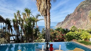 Un espace de restauration aménagé au bord de la piscine, avec des chaises et une table, offrant une vue sur un paysage montagneux et l’océan.