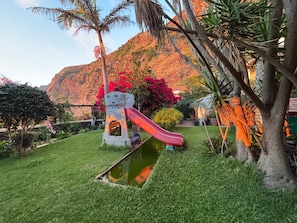 A playground with a slide and a small pool, surrounded by lush greenery and a mountain backdrop.