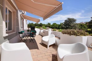 A patio with white chairs, a table, and a grill, overlooking a garden and buildings.