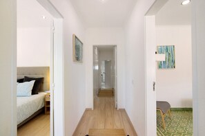 A narrow hallway with a bed on the left, a chair on the right, and a door at the end leading to another room.