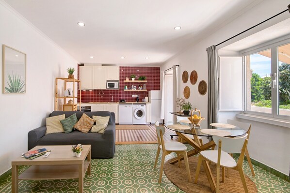 A compact living space with a sofa, coffee table, dining table, chairs, a washing machine, a microwave, and a kitchen area with red tiles and cabinets.