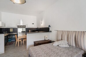 Ein kleines Zimmer mit Küchenzeile, einem Bett mit gemusterten Textilien und einem gestreiften Vorhang.