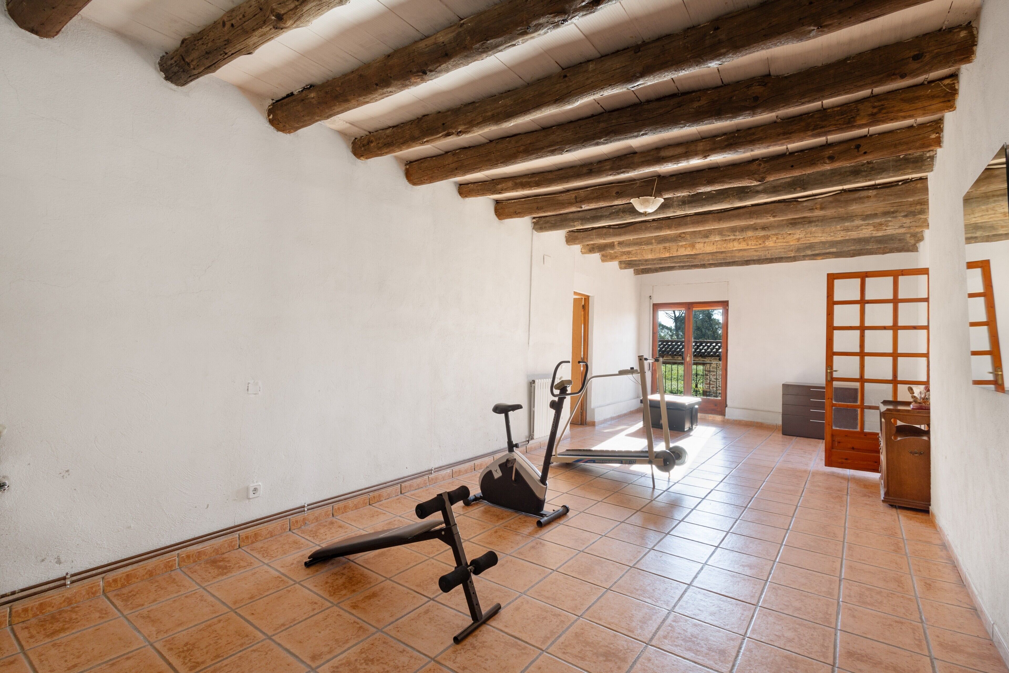 Een kamer met betegelde vloer, houten balken in het plafond, fitnessapparatuur en uitzicht door een open deur.