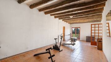 Een kamer met betegelde vloer, houten balken in het plafond, fitnessapparatuur en uitzicht door een open deur.
