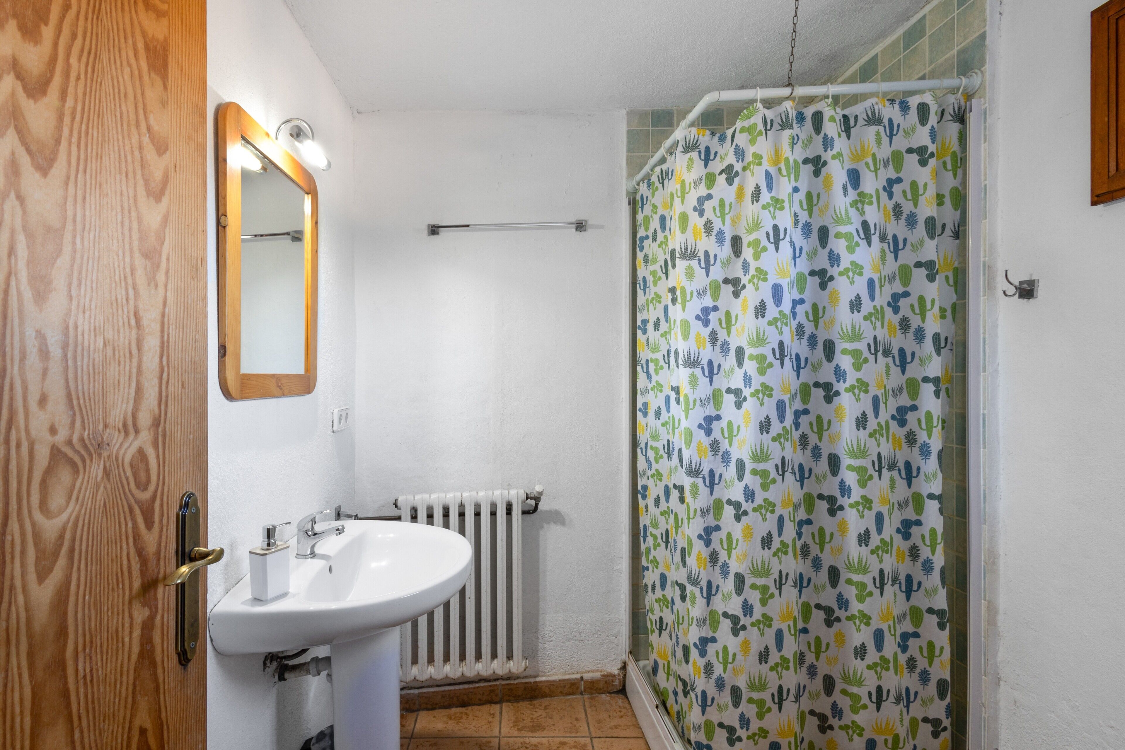 Un baño con lavamanos blanco, espejo, cortina de ducha con estampado de cactus y puerta de madera.