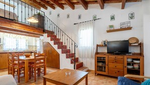 A cozy living room with a wooden dining table, a sofa, a TV on a wooden cabinet, and a staircase leading to an upper level. - Country House 'Caseta Montanyana' with Private Terrace, Private Garden and Wi-Fi (Deltebre)