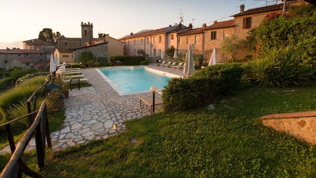 A pool area with stone flooring, lounge chairs, and umbrellas, surrounded by stone buildings and greenery. - Apartment 'Appartamento Paggio' with Shared Pool and Air Conditioning (Massarosa)