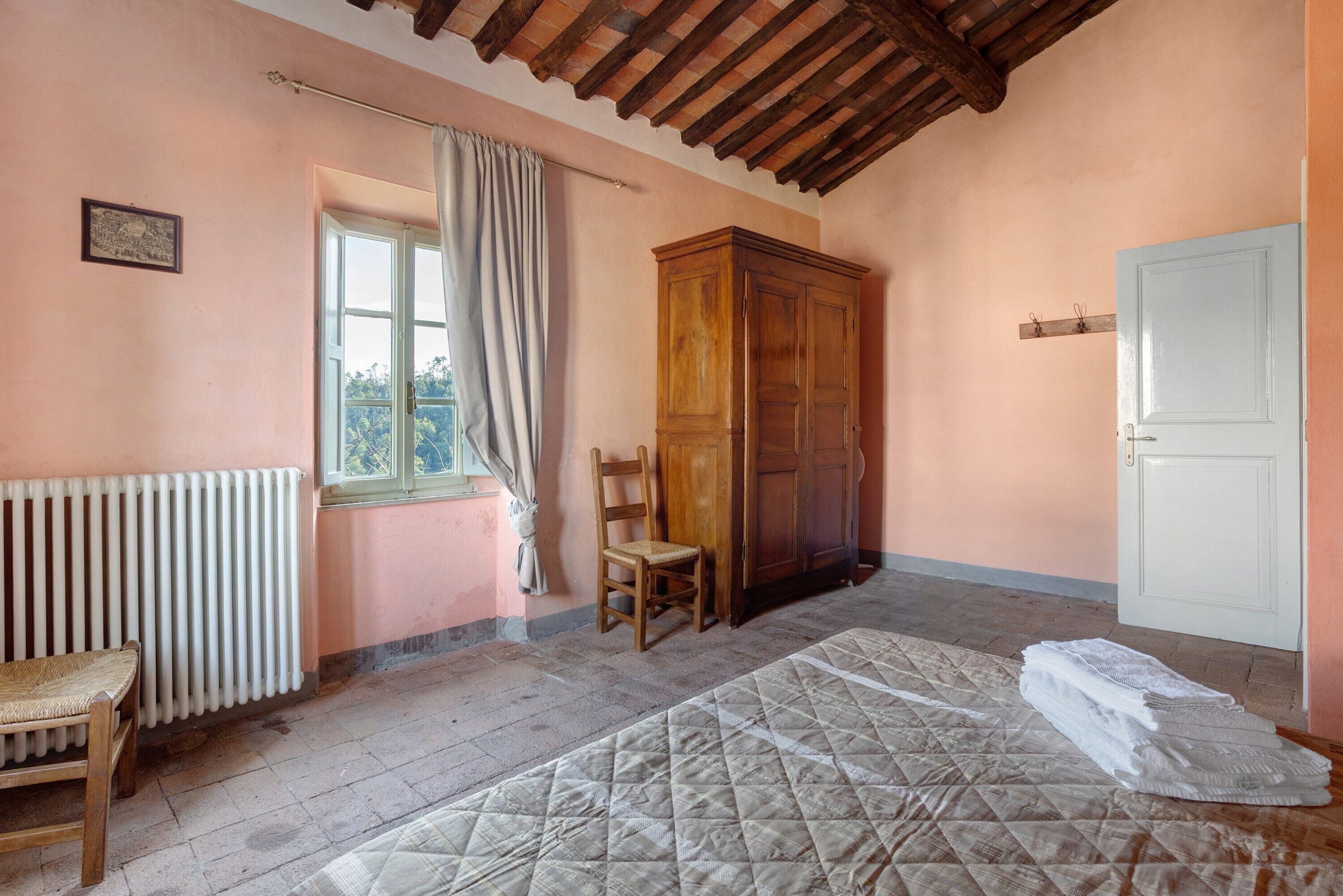 Quarto com radiador, cadeira de madeira, cama com colcha, guarda-roupa de madeira, janela com cortinas e porta branca.