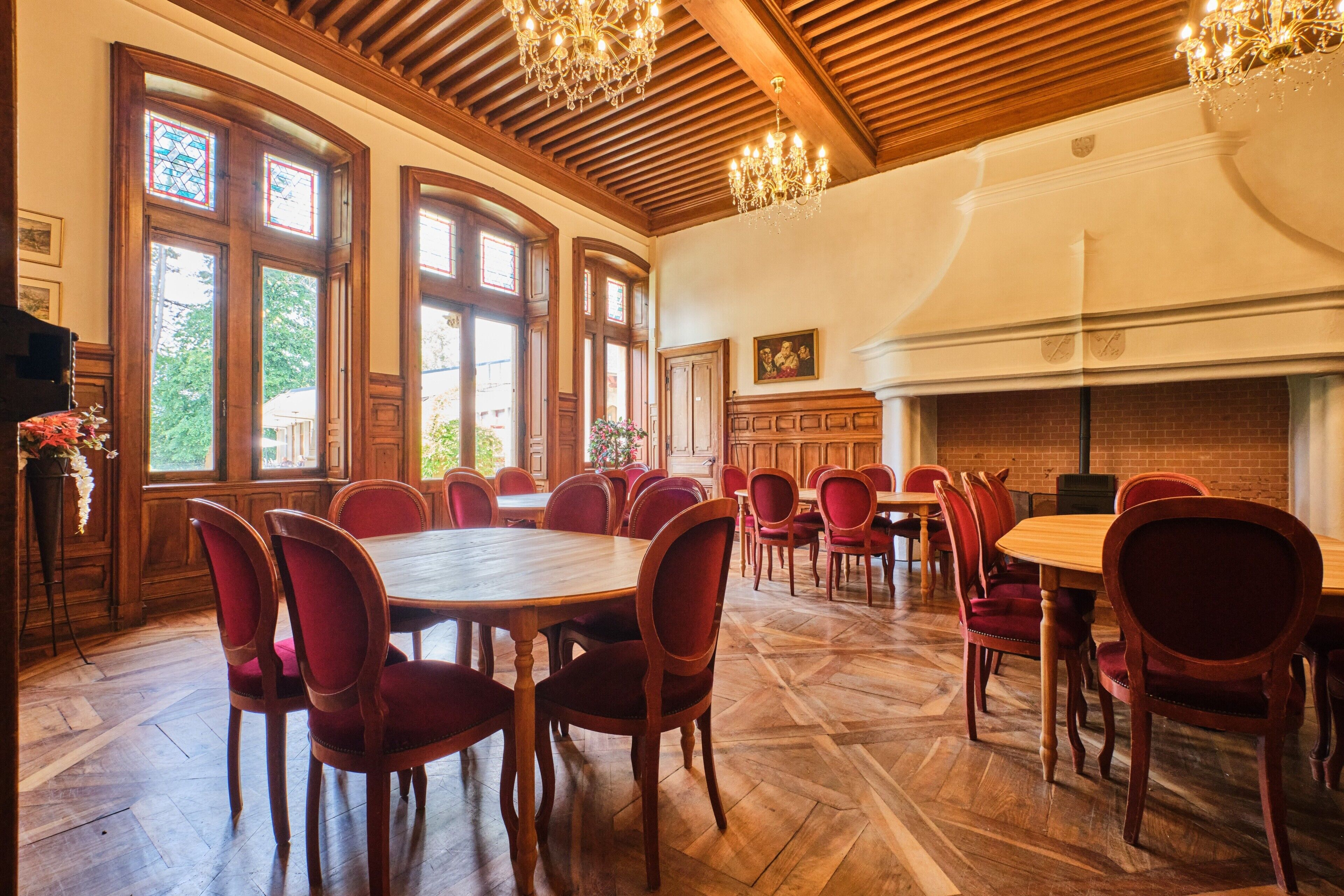 Un espace repas avec des tables en bois et des chaises rouges, de grandes fenêtres et une cheminée en briques.