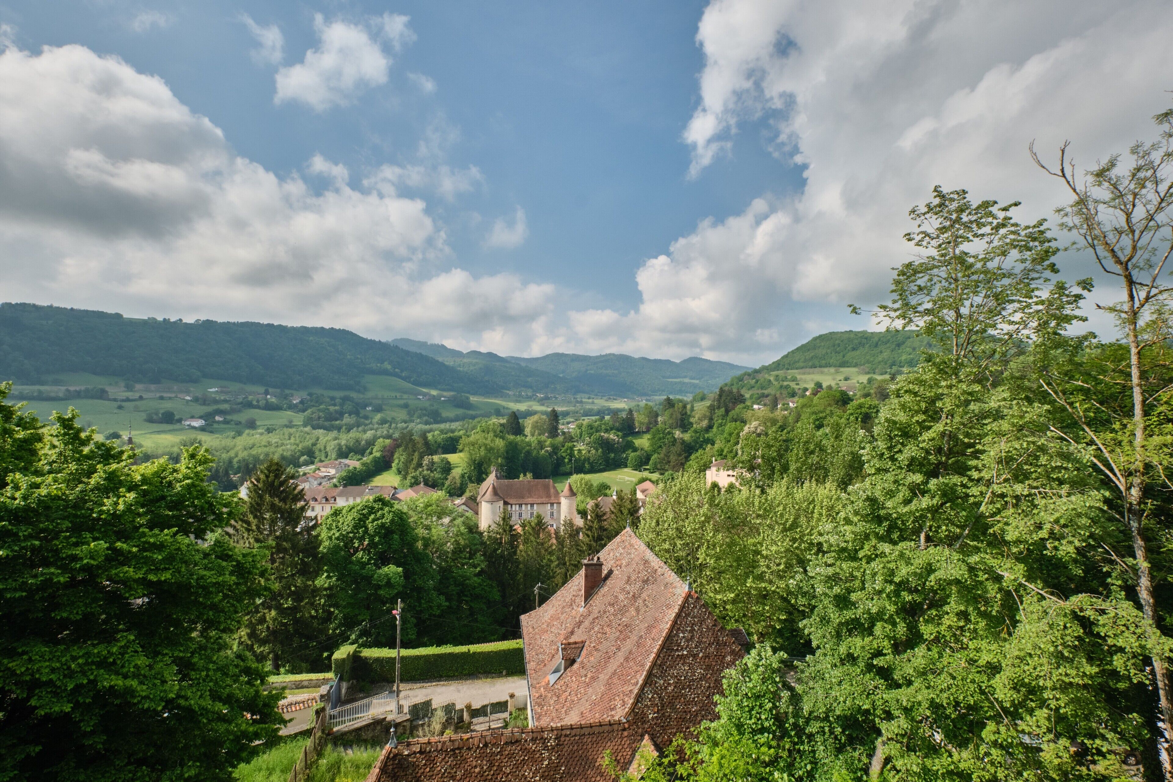 Un paysage rural dominé par un bâtiment imposant, doté d’un toit en tuiles, entouré d’une végétation luxuriante et bordé de collines lointaines.