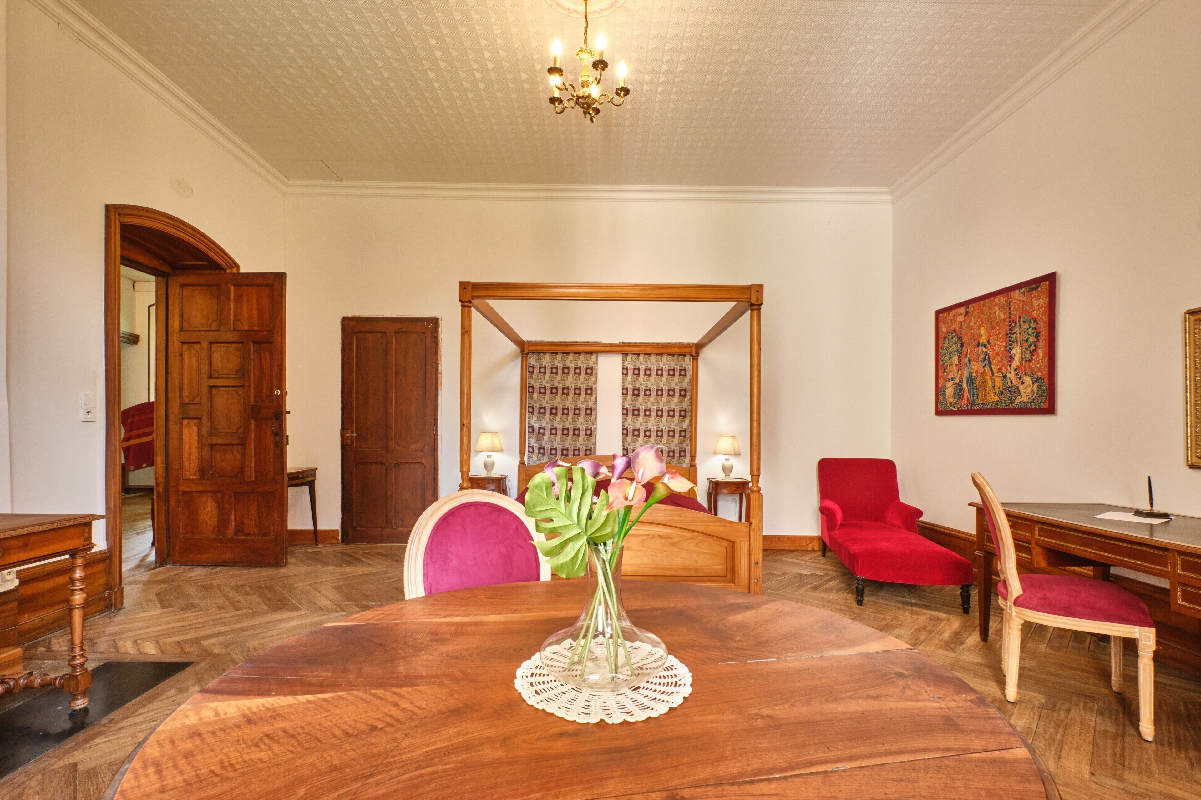 Ein Zimmer mit einem hölzernen Himmelbett, einem Esstisch mit Blumenvase, einem roten Sessel und einem Schreibtisch mit Stuhl.