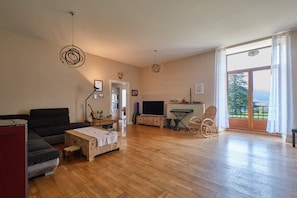 A spacious living room with a wooden floor, a sofa, a television, and a view of a balcony. - Guest House 'Suite Marguerite Chez L'habitant' with Mountain View, Shared Terrace and Shared Garden (Saint-Geoire-en-Valdaine)
