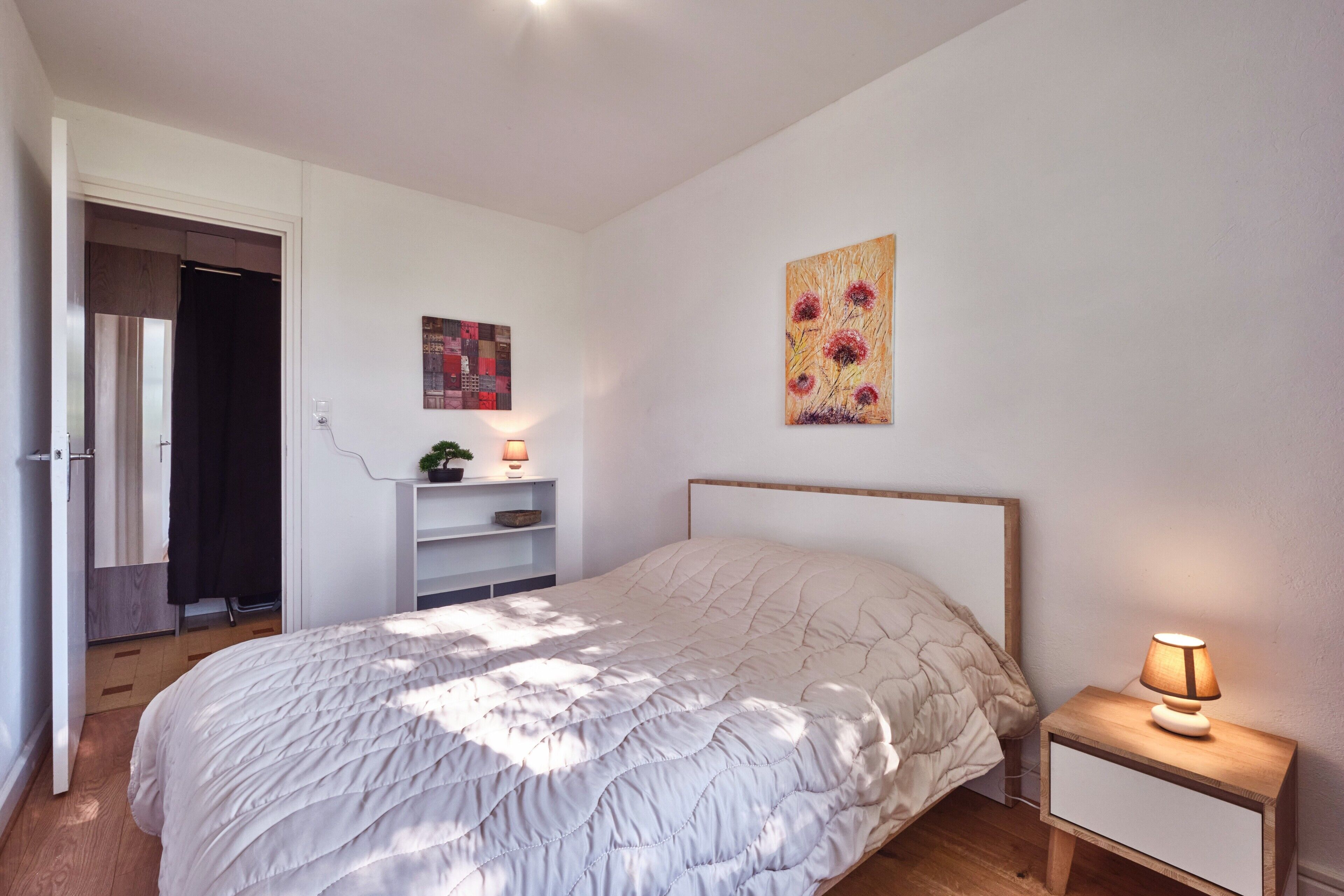 Une chambre à coucher comprenant un lit, une table de chevet, une lampe, des œuvres d’art murales et une porte donnant sur une autre pièce.