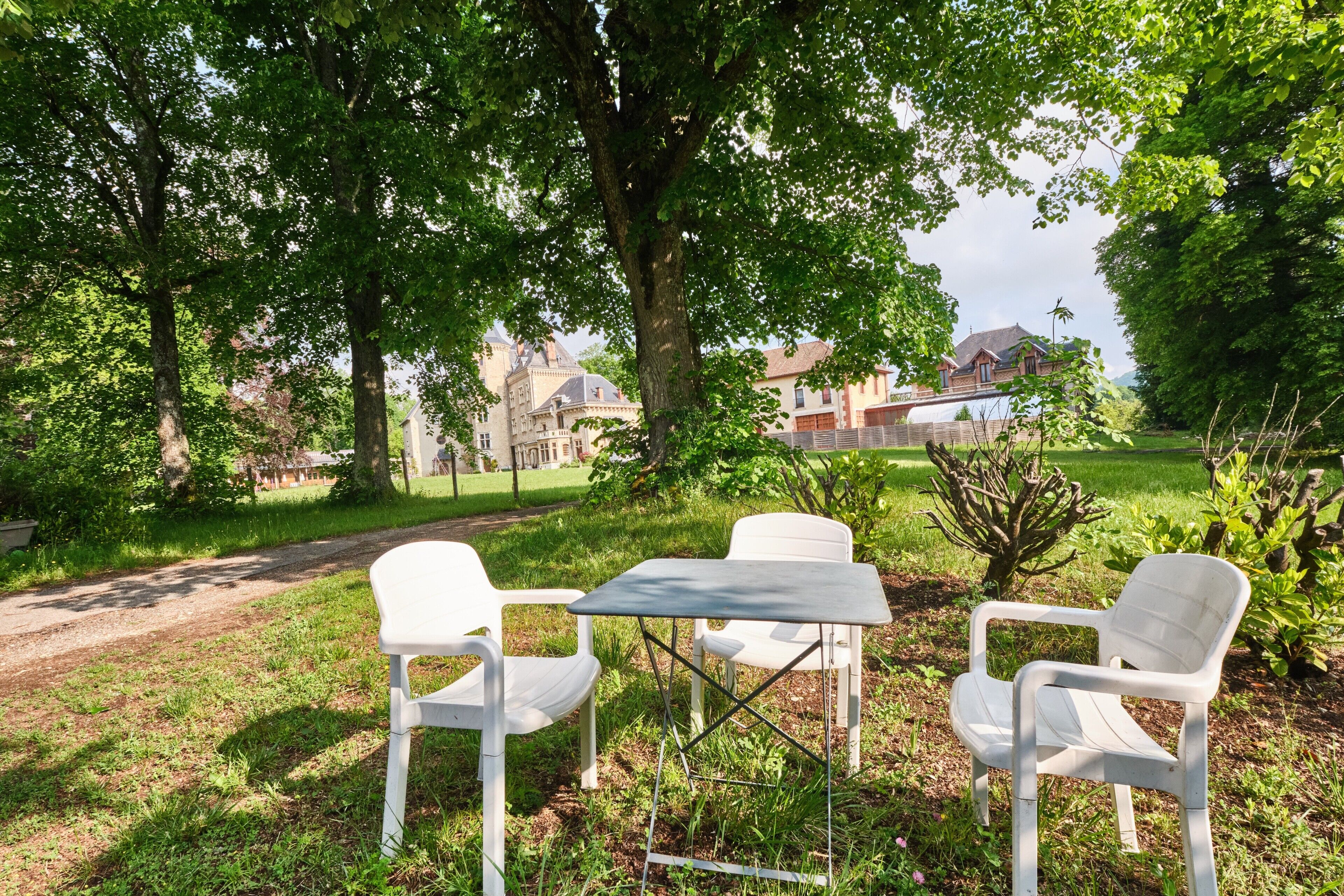 Un área de estar al aire libre con sillas de plástico blanco y una mesa, rodeada de árboles y un césped.