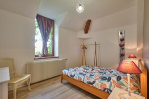 A bedroom with a bed, a chair, a radiator, a window with curtains, a lamp, and a wooden artwork on the wall. - Apartment 'Grand Gîte Du Château De St Geoire' with Mountain View, Private Terrace and Shared Garden (Saint-Geoire-en-Valdaine)
