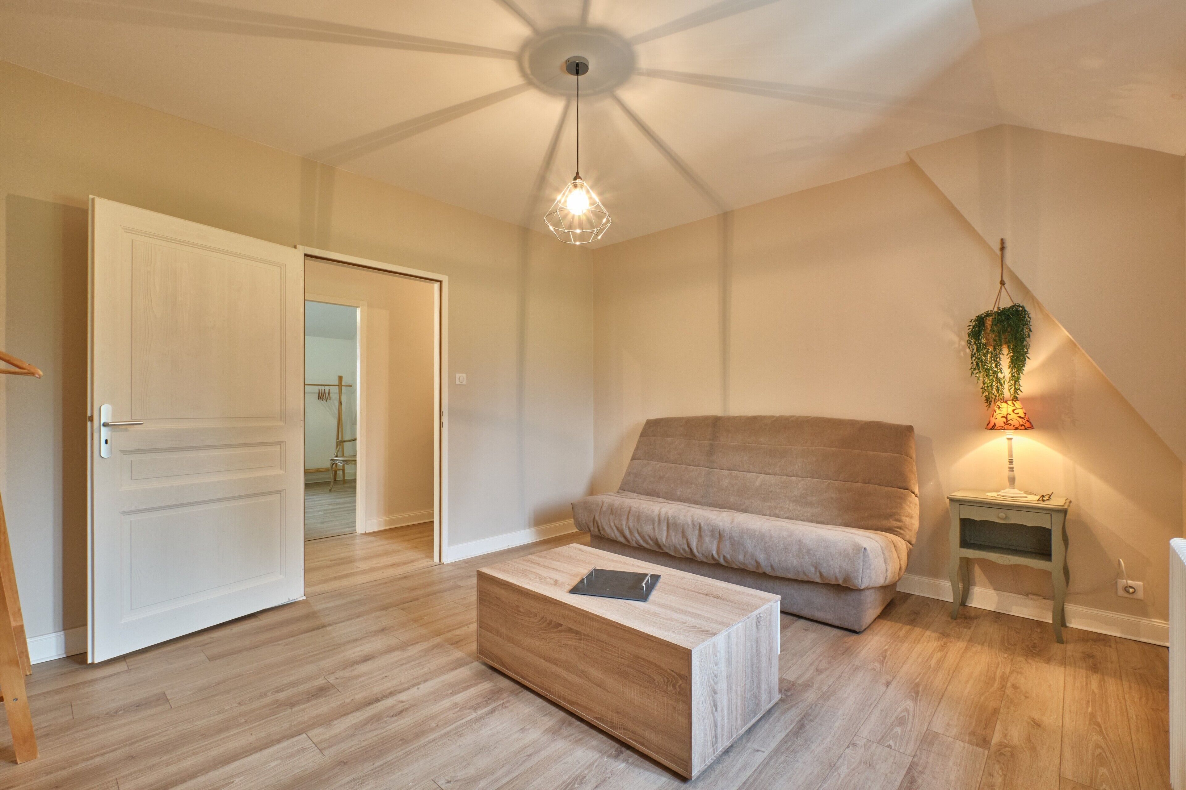 A room with a wooden floor, a sofa, a coffee table, a hanging light fixture, and a small bedside table with a lamp.