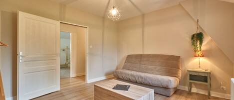 A room with a wooden floor, a sofa, a coffee table, a hanging light fixture, and a small bedside table with a lamp.