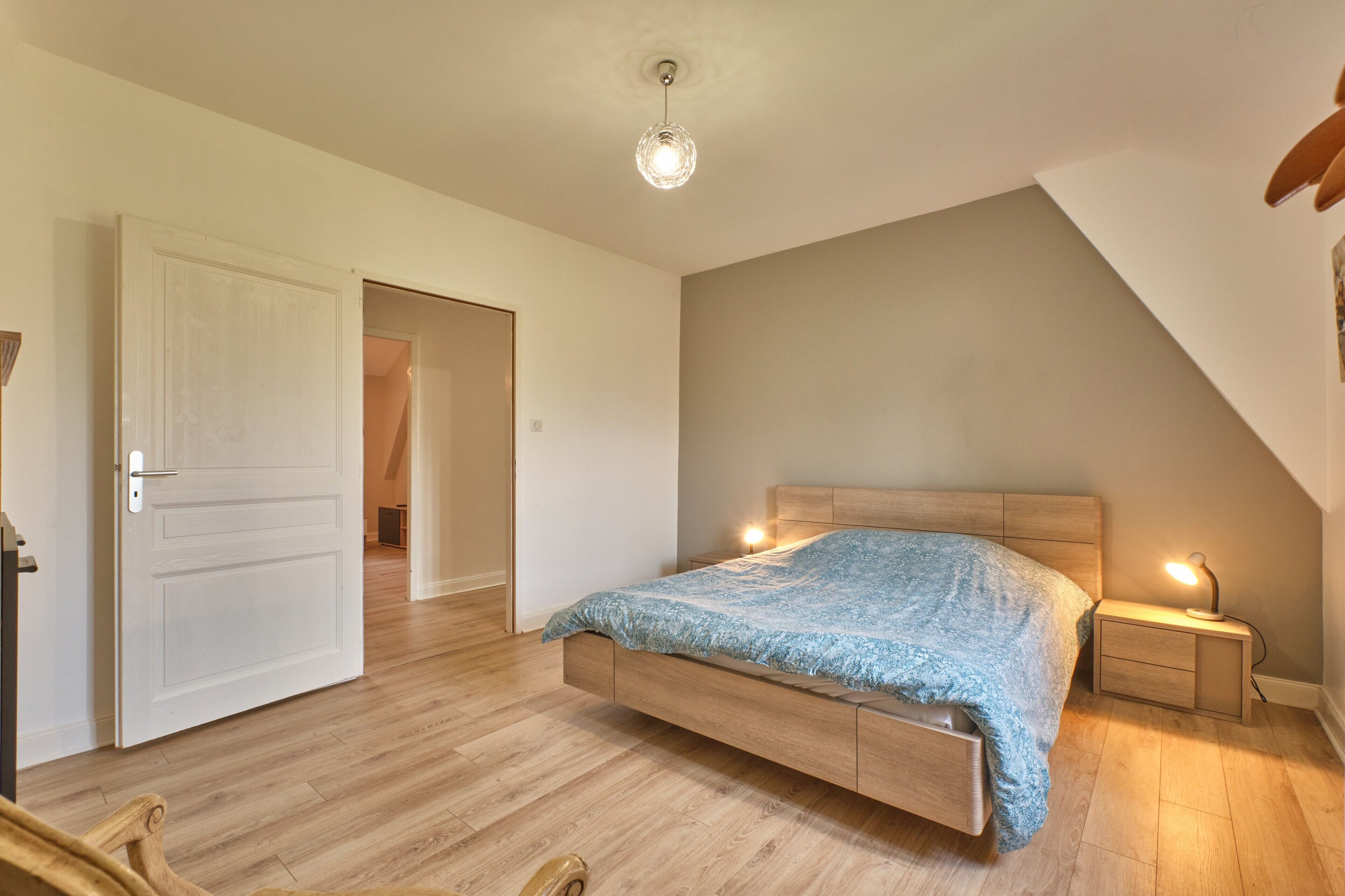 A bedroom with a wooden bed, bedside tables, a ceiling light, and a door leading to another room.