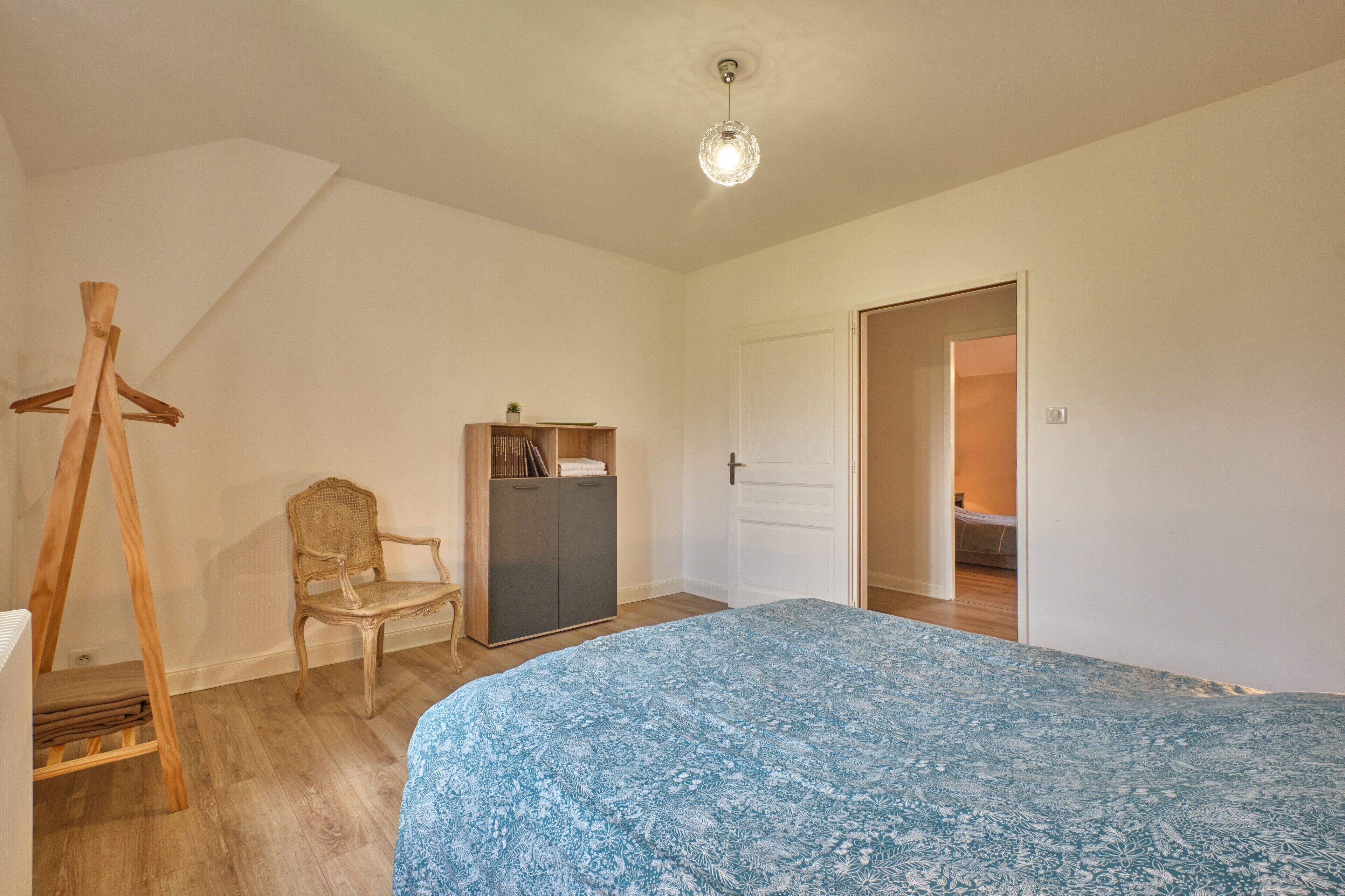 A bedroom with a bed, a wooden chair, a wardrobe, and a door leading to another room.