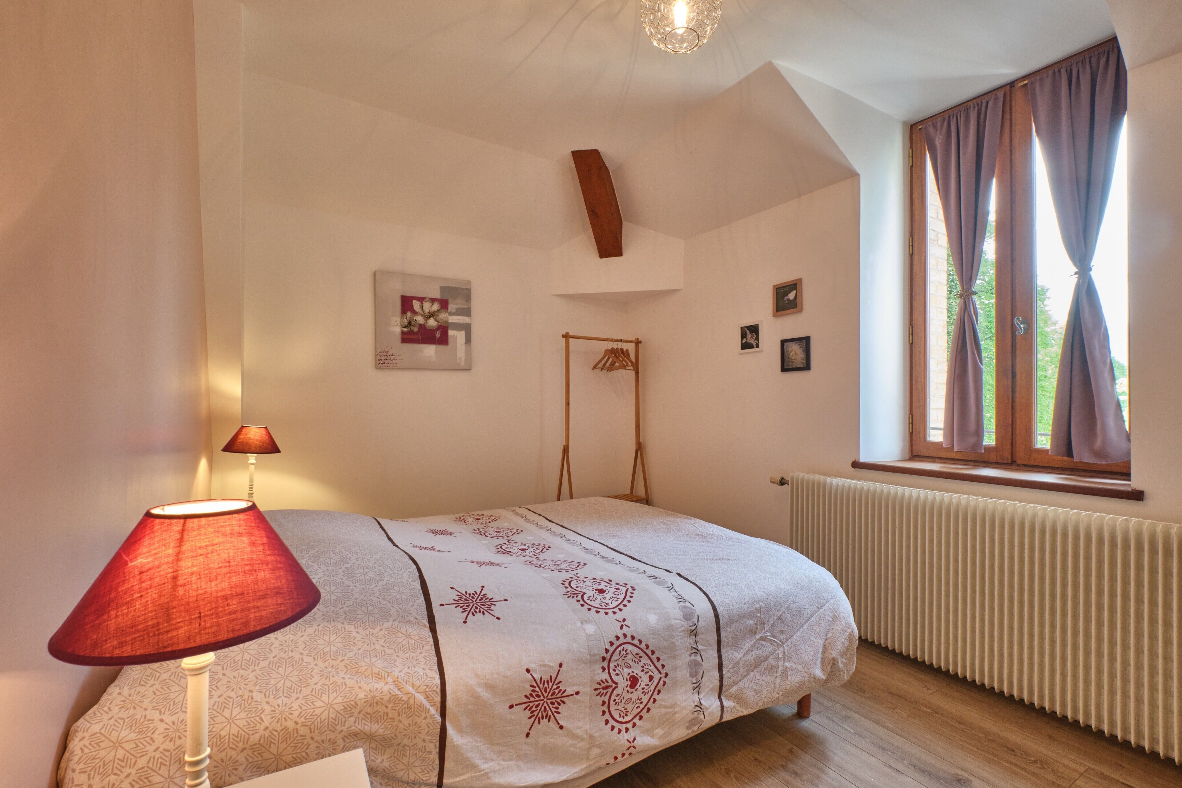 A bedroom with a bed, a lamp, a window with curtains, a radiator, and wall-mounted pictures.