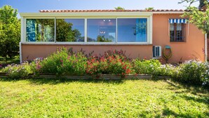 Una casa con un ventanal grande, una ventana pequeña con toldo azul, y un jardín con diversas plantas y flores.