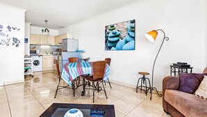 A modern living room with a sofa, a coffee table, a dining area with chairs and a table, a kitchen in the background, and a wall-mounted lamp. - Apartment 'Superbe Appartement Vue Mer' with Sea View, Wi-Fi and Air Conditioning (Fréjus)