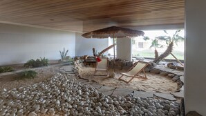 A covered outdoor area with a wooden ceiling, a thatched umbrella, two lounge chairs, and a sandy ground with rocks. - Apartment 'Superbe Appartement Vue Mer' with Sea View, Wi-Fi and Air Conditioning (Fréjus)