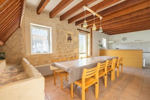 A dining area with a long table, wooden chairs, stone walls, and a kitchen in the background. - Charming House 'Grands Gites 16 Personnes' with Private Terrace, Garden & Wi-Fi (La Gripperie-Saint-Symphorien)