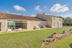 A stone house with a tiled roof, a glass door leading to a patio with chairs and a pool, and a stone wall with planters. - Charming House 'Grands Gites 16 Personnes' with Private Terrace, Garden & Wi-Fi (La Gripperie-Saint-Symphorien)
