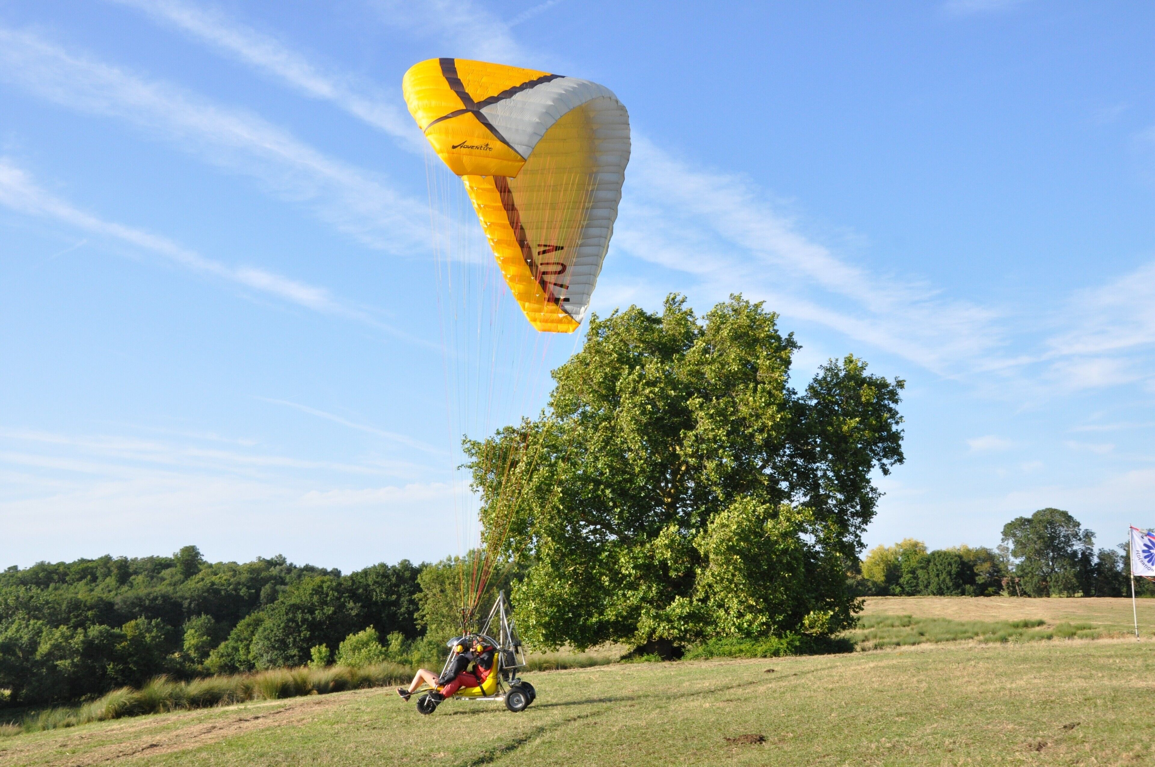 Una persona practicando parapente con un paracaídas sobre un campo cubierto de hierba, con árboles y una bandera al fondo.