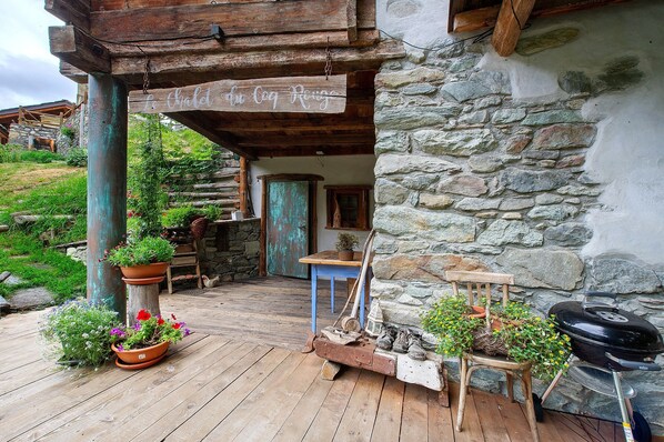 A rustic wooden porch with a stone wall, a wooden chair, a table, and a grill.