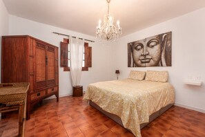 Une chambre à coucher avec un lit, une armoire en bois, un lustre, un tableau mural et une fenêtre avec des rideaux.
