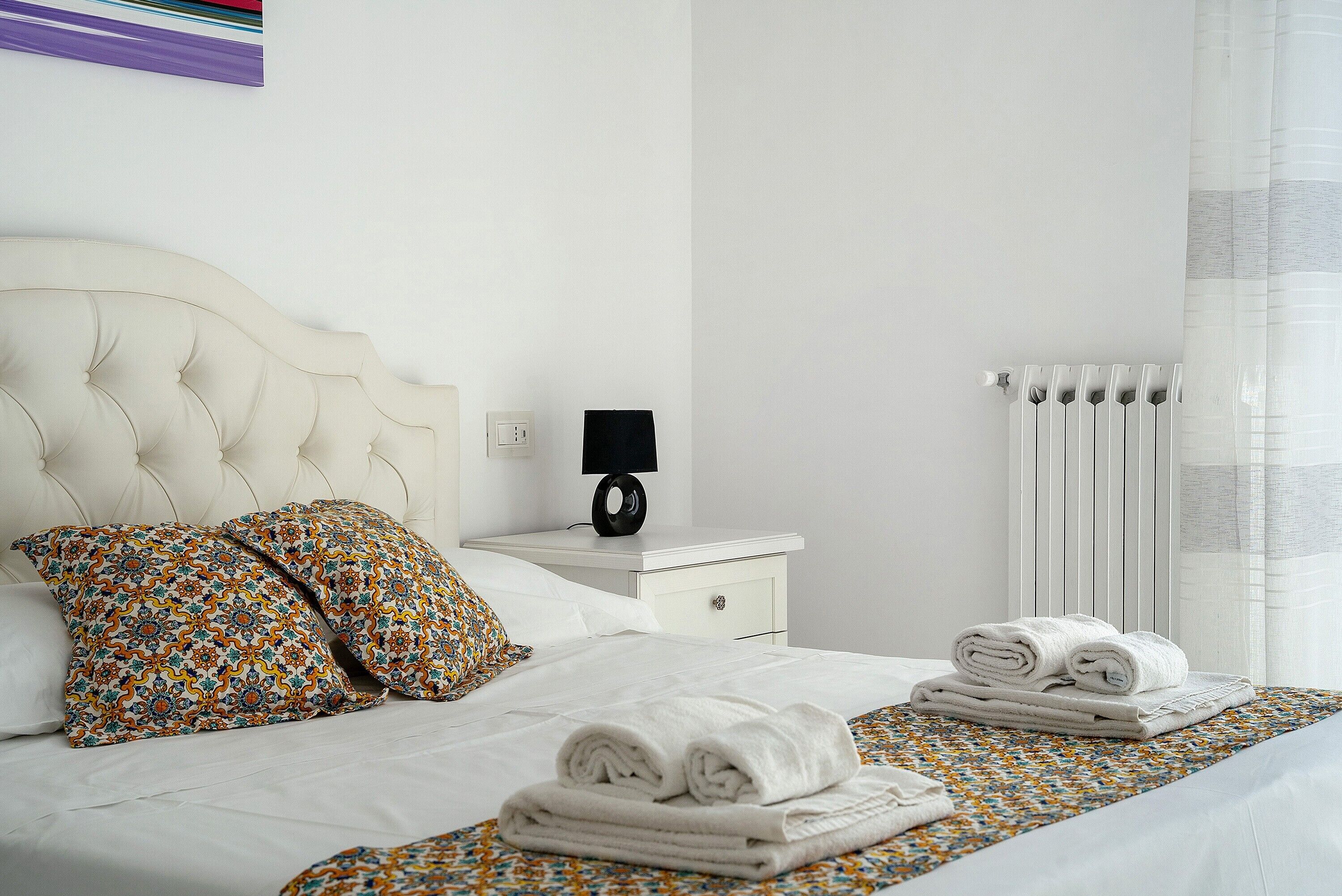A neatly made bed with white linens, a patterned pillow, and rolled towels on a bed with a patterned bedspread.