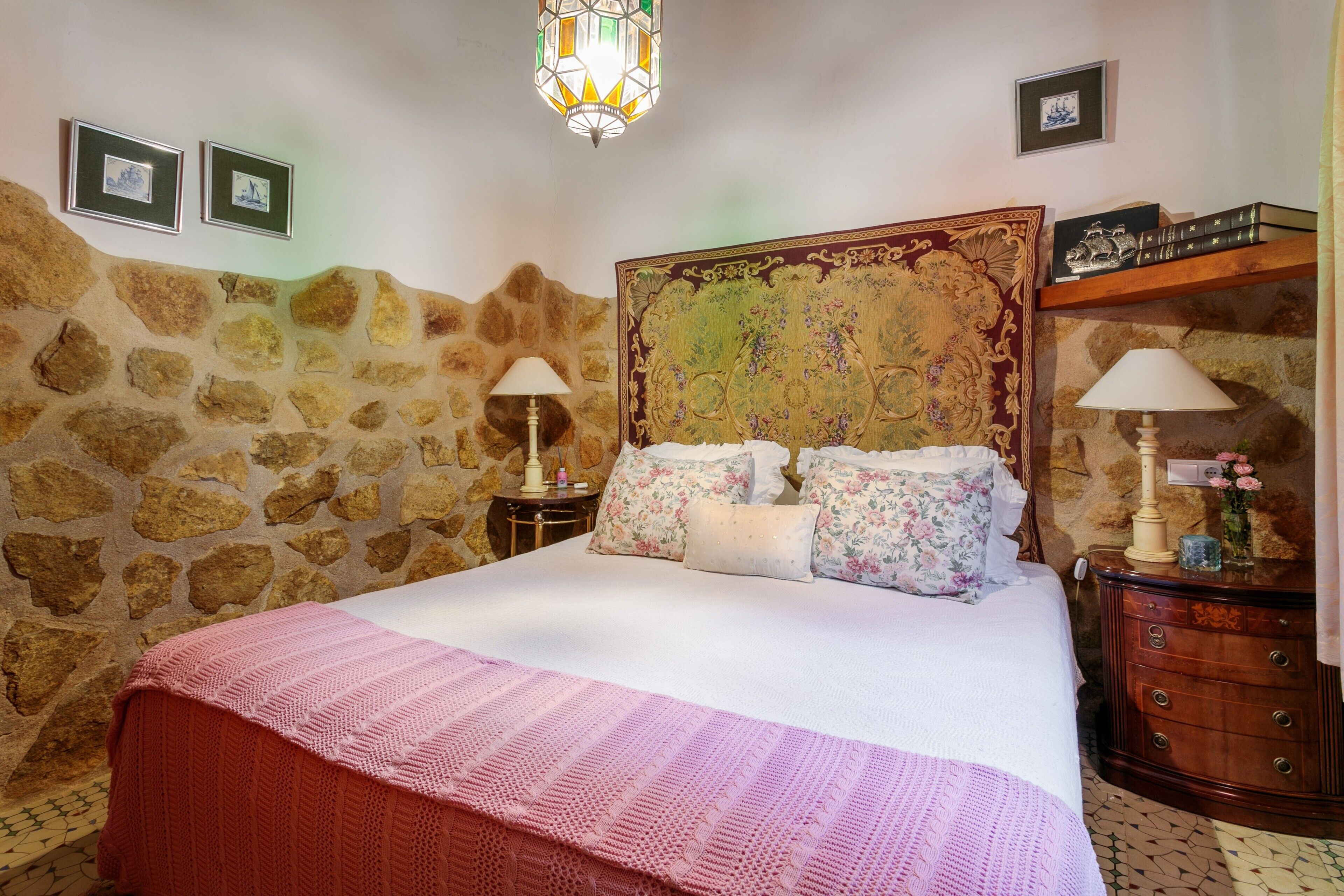 Un dormitorio con pared de piedra, una cama grande con almohadas florales, un aparador de madera y una araña.