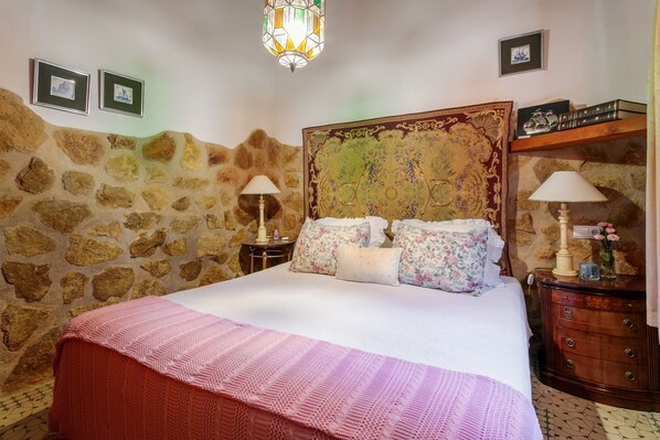 Une chambre avec un mur en pierre, un grand lit agrémenté d’oreillers à motifs floraux, une commode en bois et un lustre.