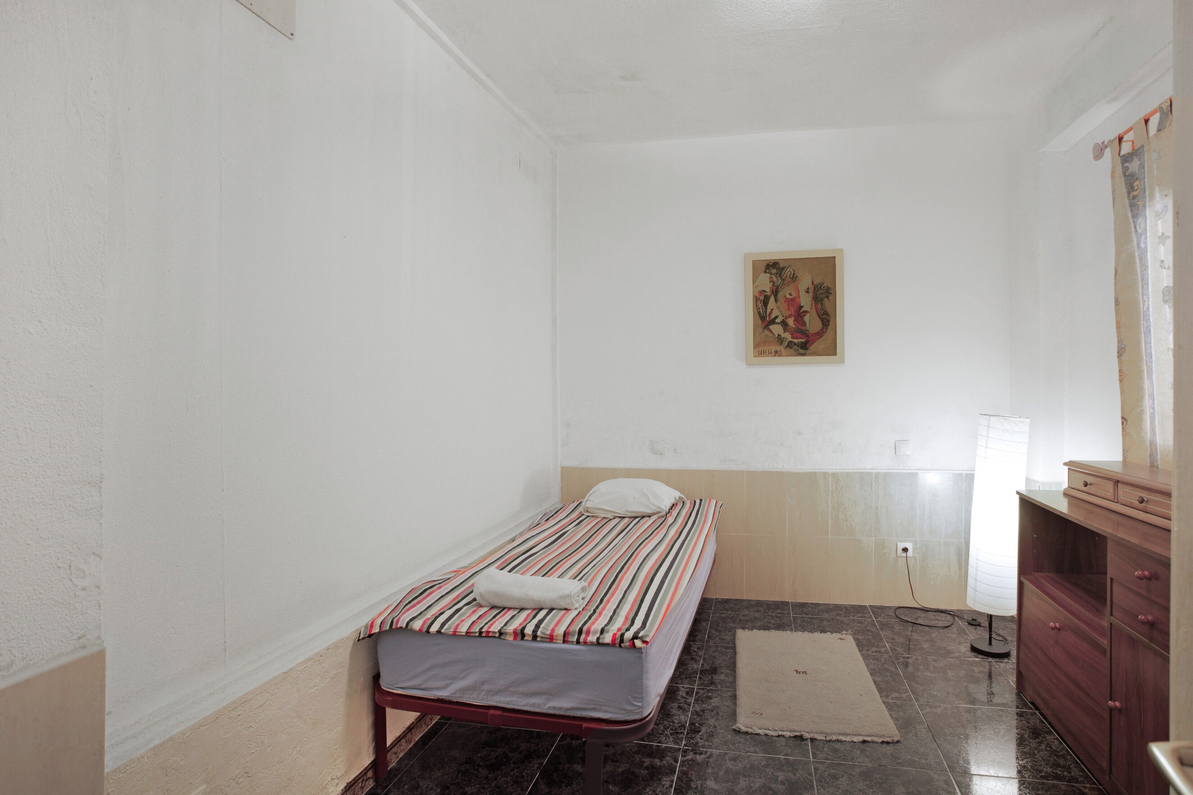 Una stanza piccola con un letto singolo, una coperta a righe, un armadietto in legno, un quadro appeso al muro e un pavimento piastrellato.