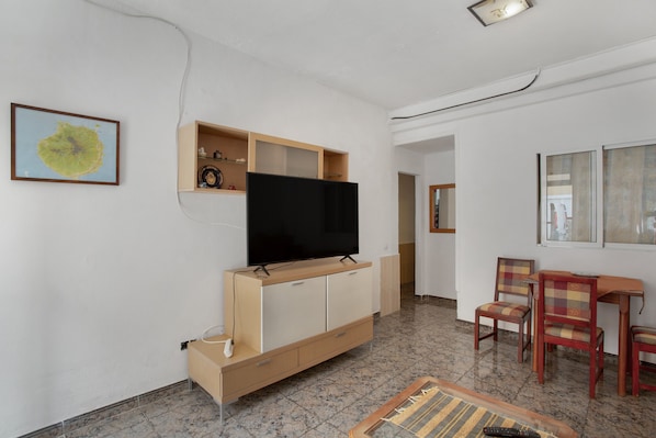 A living room with a flat-screen TV, wooden cabinets, a coffee table, and a dining area with chairs.
