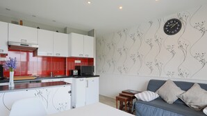 Une cuisine moderne avec des meubles blancs et des touches de rouge, un coin repas avec une table blanche, et un canapé bleu agrémenté de coussins.